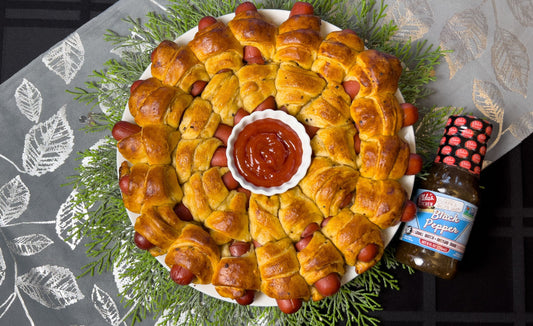 Pigs in a blanket arranged in a wreath shape with a bottle of Elda's Kitchen Black Pepper sauce.