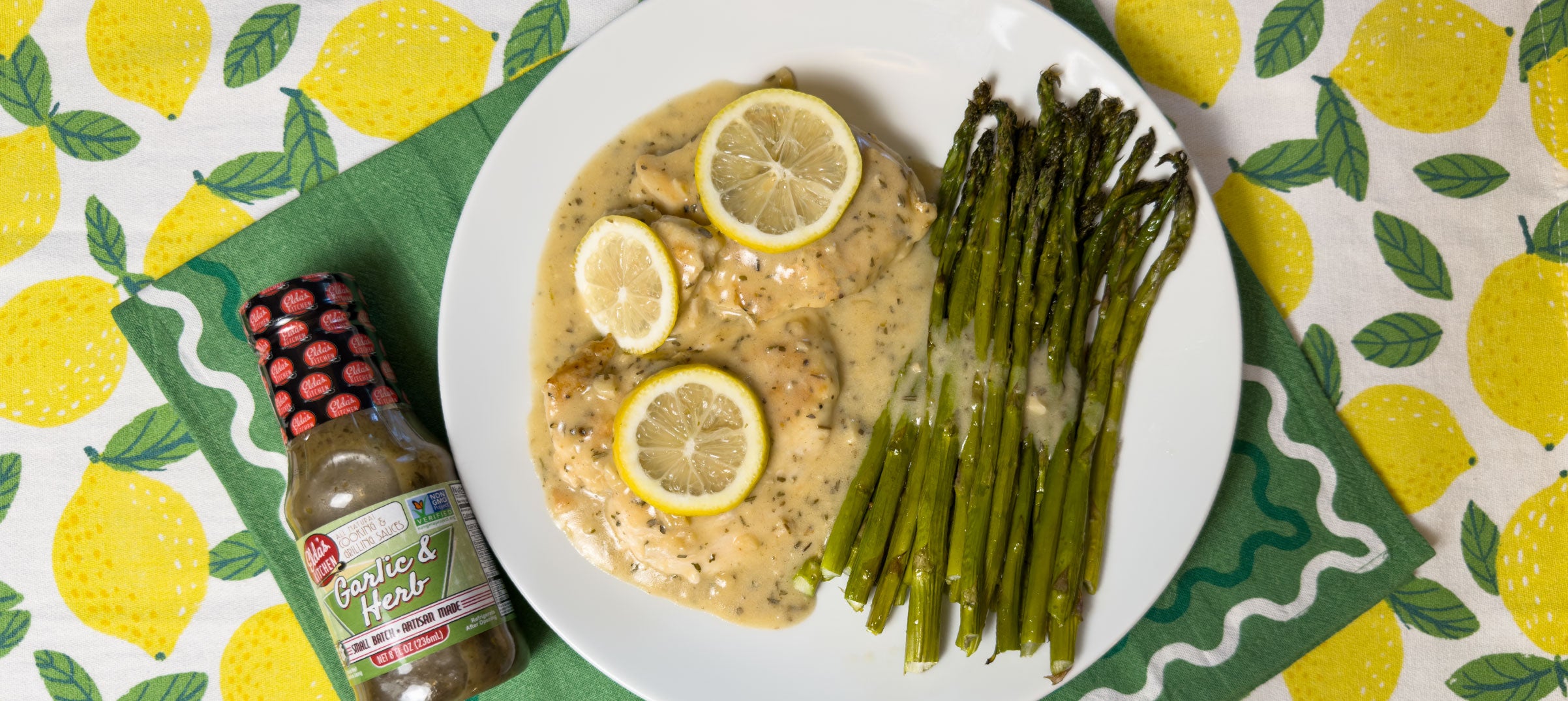 Plate of lemon garlic chicken and asparagus with a bottle of Elda's Kitchen Garlic & Herb Sauce.