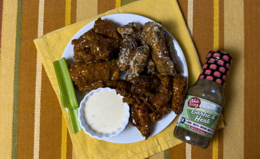 A plate of baked chicken wings with celery and ranch dressing next to a bottle of Elda's Kitchen honey Ginger & Soy Sauce.