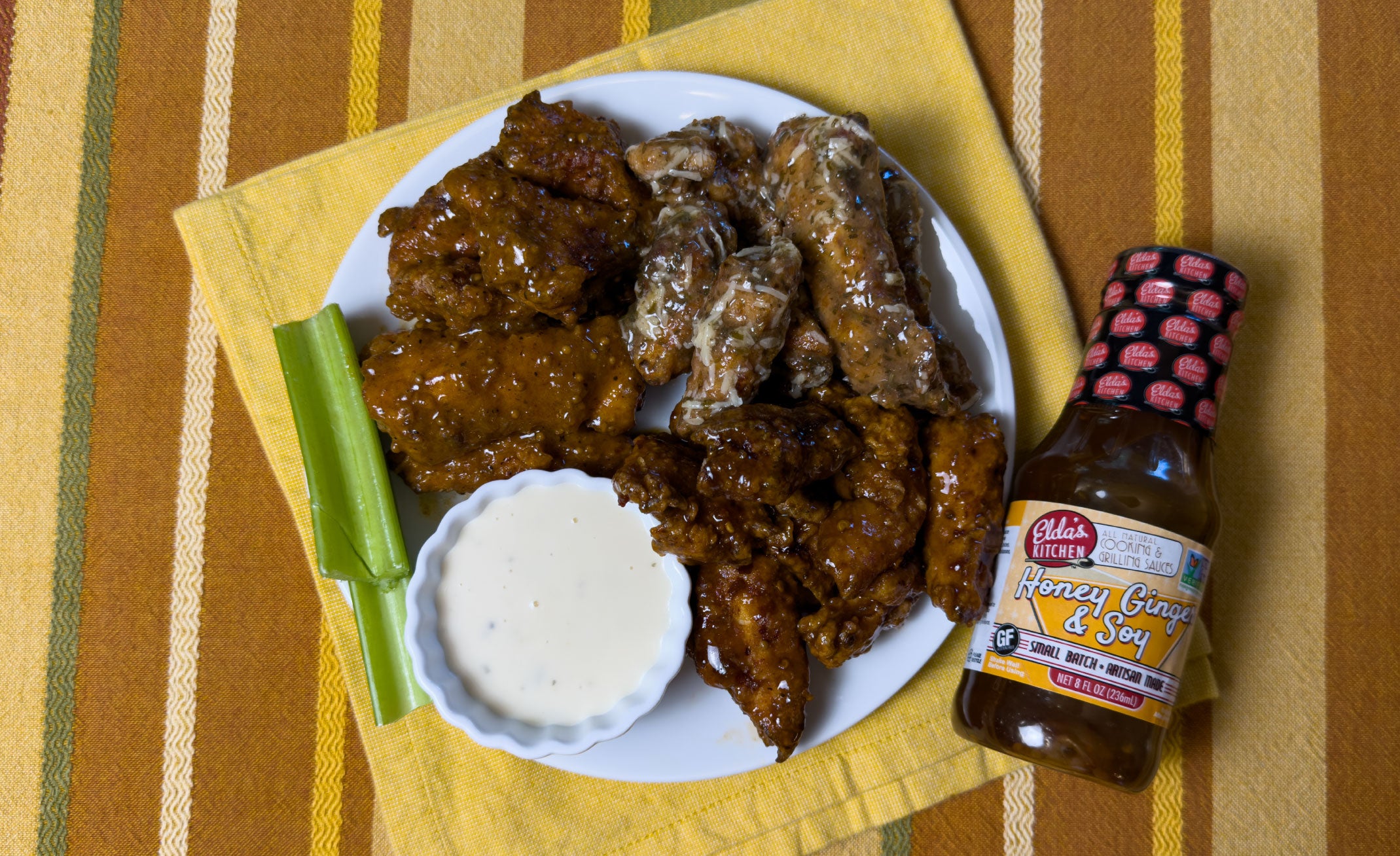 A plate of baked chicken wings with celery and ranch dressing next to a bottle of Elda's Kitchen honey Ginger & Soy Sauce.
