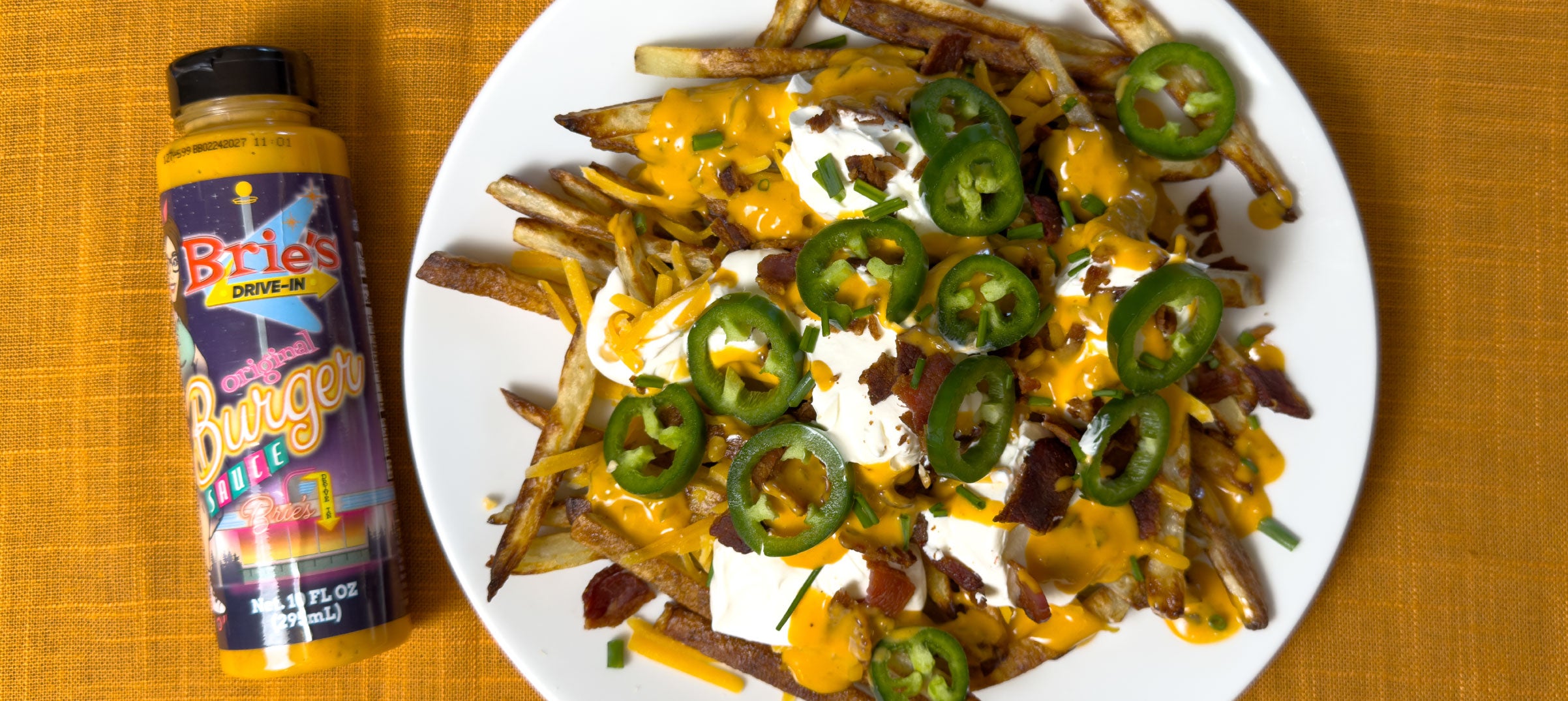 Plate of loaded french fries with sour cream and fresh jalapenos next to a bottle of Brie's Drive-In Burger Sauce.