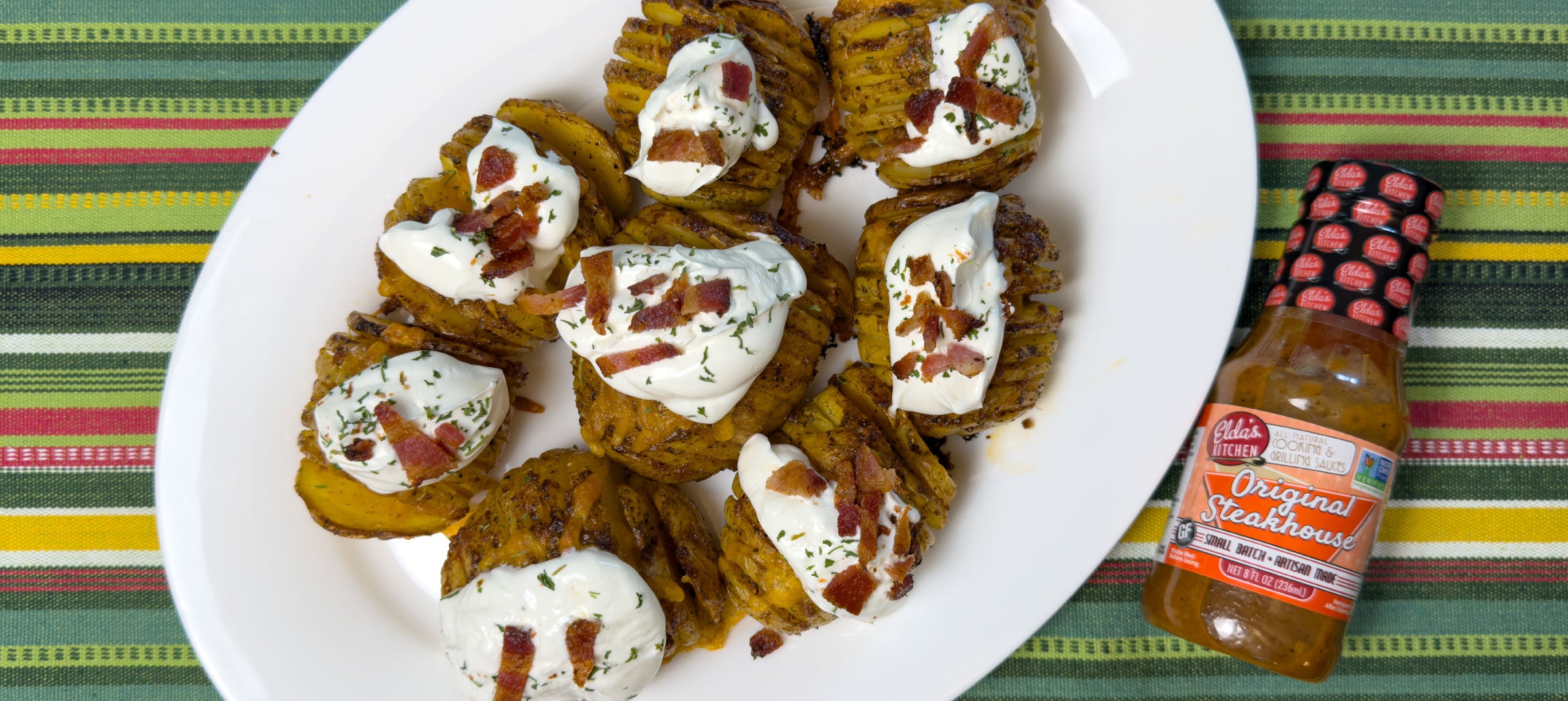 Plate of loaded, sliced baked potatoes topped with sourcream and bacon, next to a bottle of Elda's Kitchen Original Steakhouse Sauce.