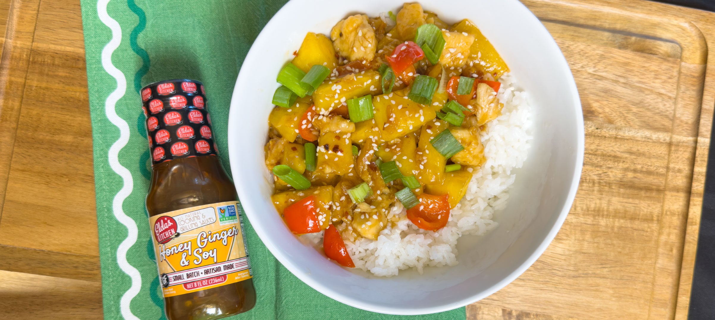 A bowl of rice with chicken and pineapple next to a bottle of Elda's Kitchen Honey Ginger & Soy Sauce