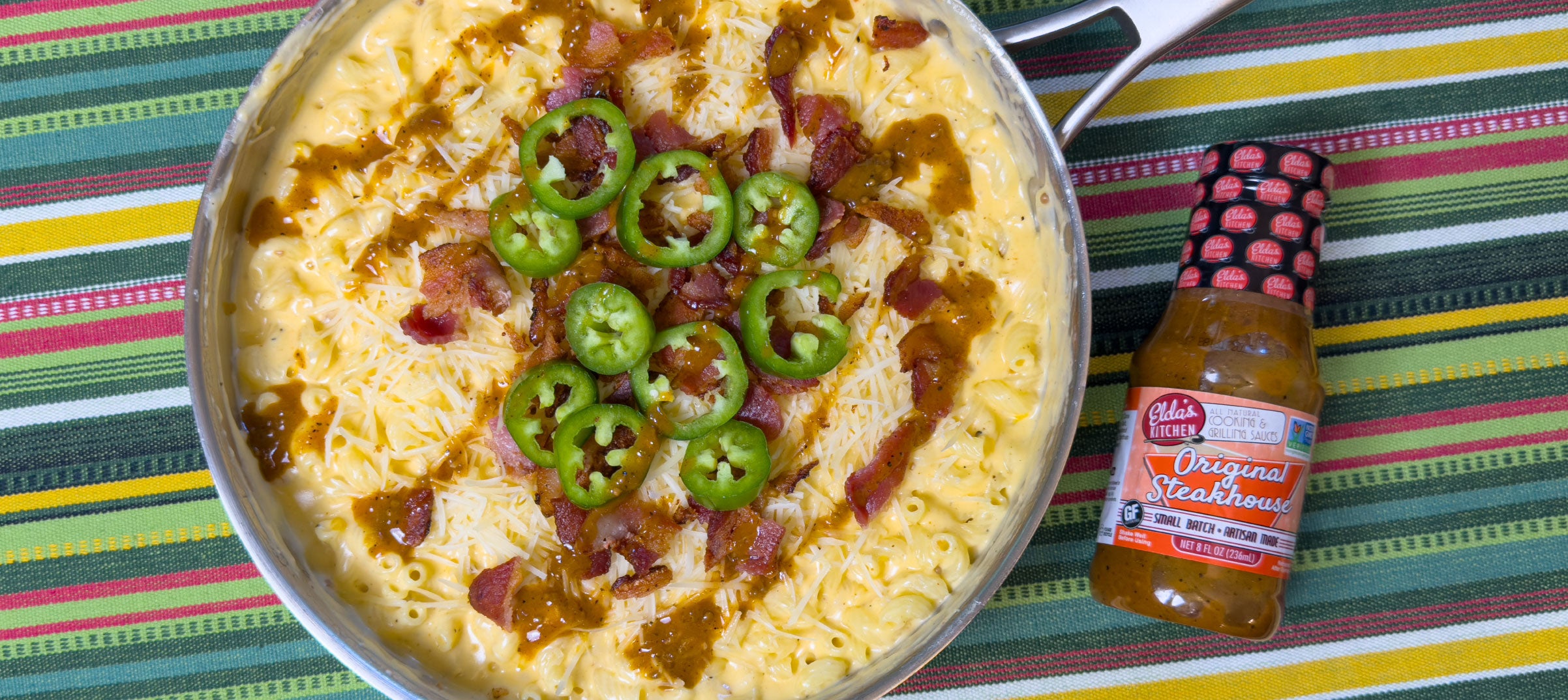 Skillet of macaroni and cheese topped with bacon and jalapeno slices, next to a bottle of Elda's Kitchen Original Steakhouse Sauce.